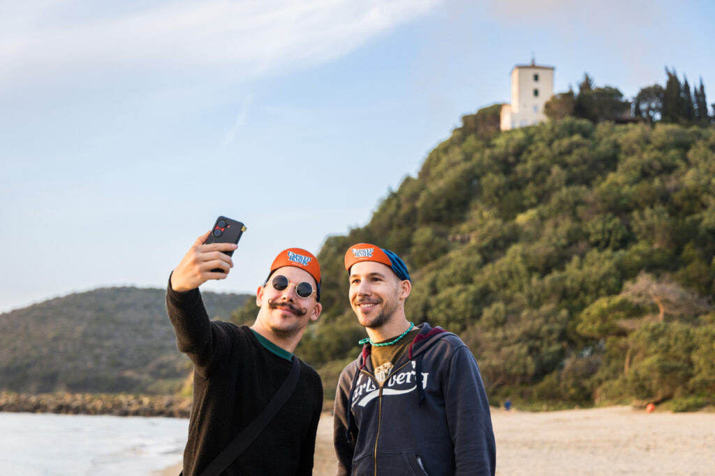Partecipanti di Unpaved Roads Punta Ala scattano un selfie in spiaggia con la Torre di Cala Violina sullo sfondo