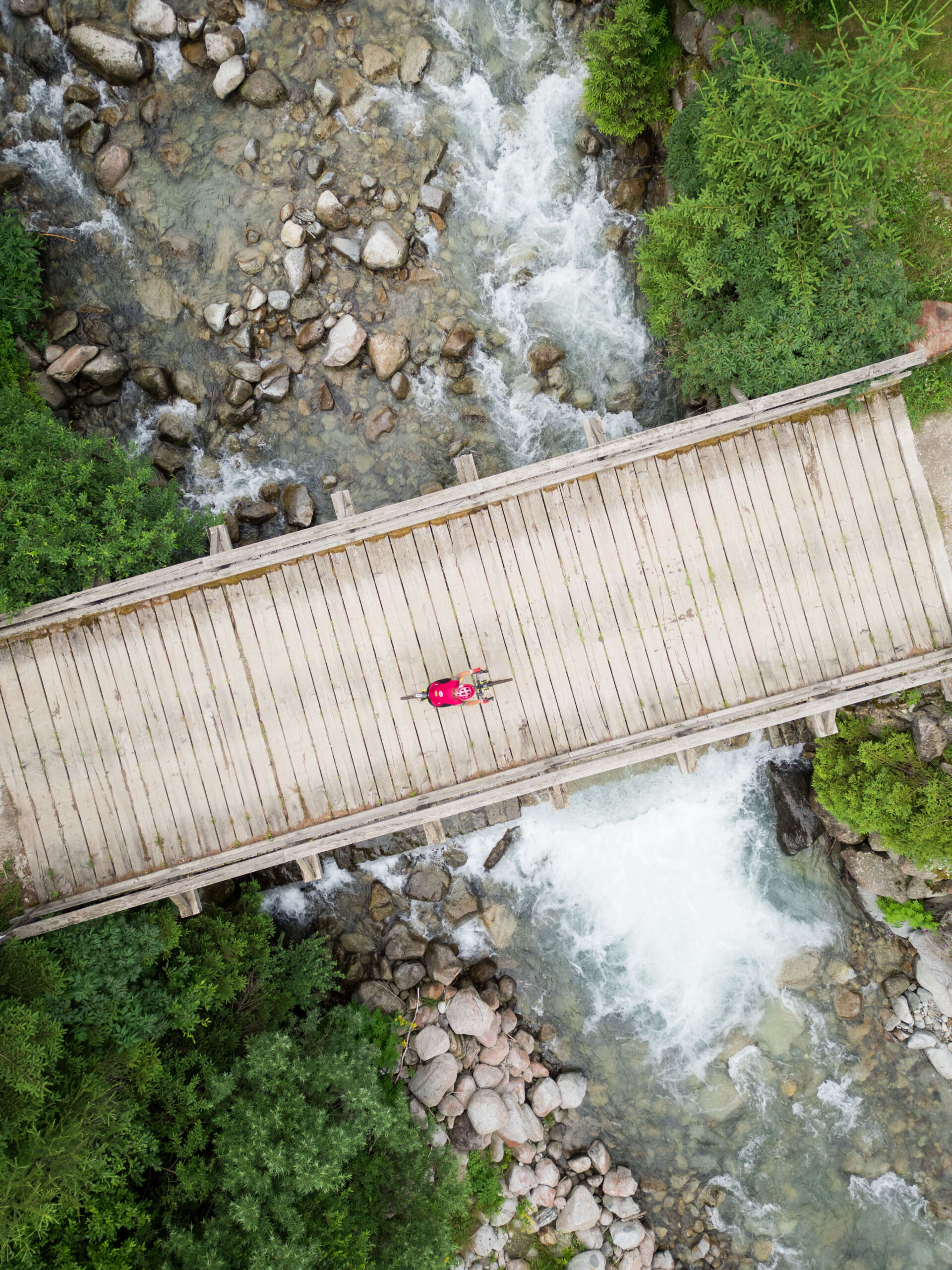 Vista aerea verticale di un ciclista che attraversa un suggestivo ponte di legno sopra un torrente di montagna durante l'Unpaved Roads