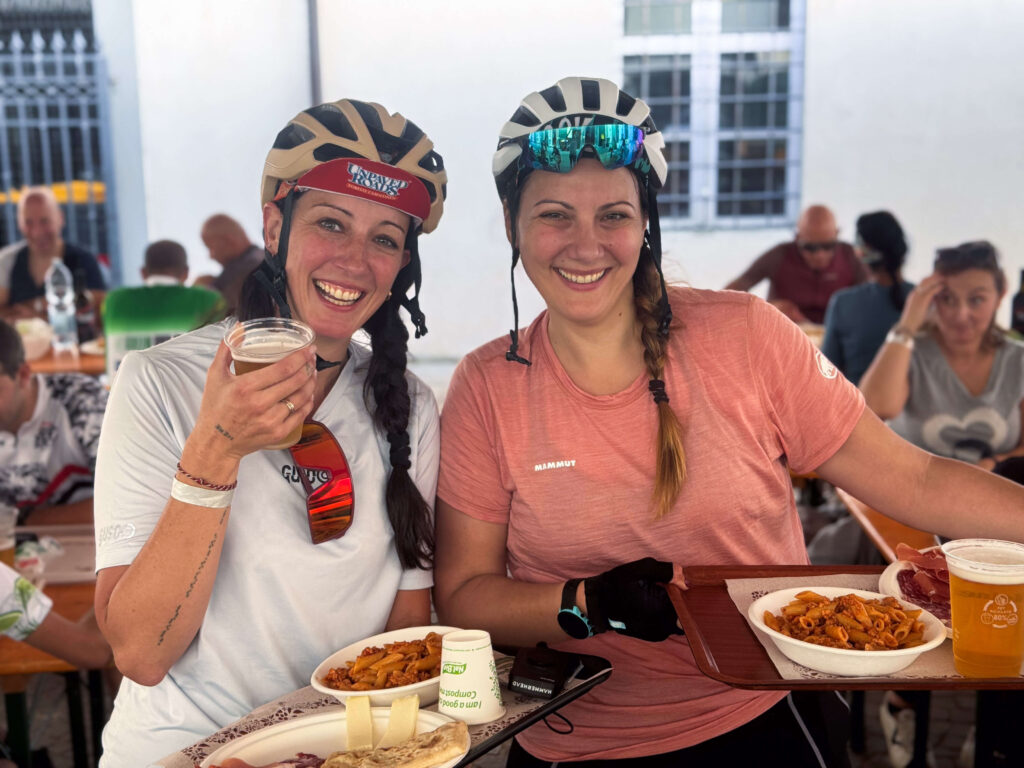 Due cicliste sorridenti festeggiano il traguardo dell'evento Unpaved Roads Foreste Casentinesi a Santa Sofia, godendosi un piatto di pasta tipica e una birra al ristoro finale.