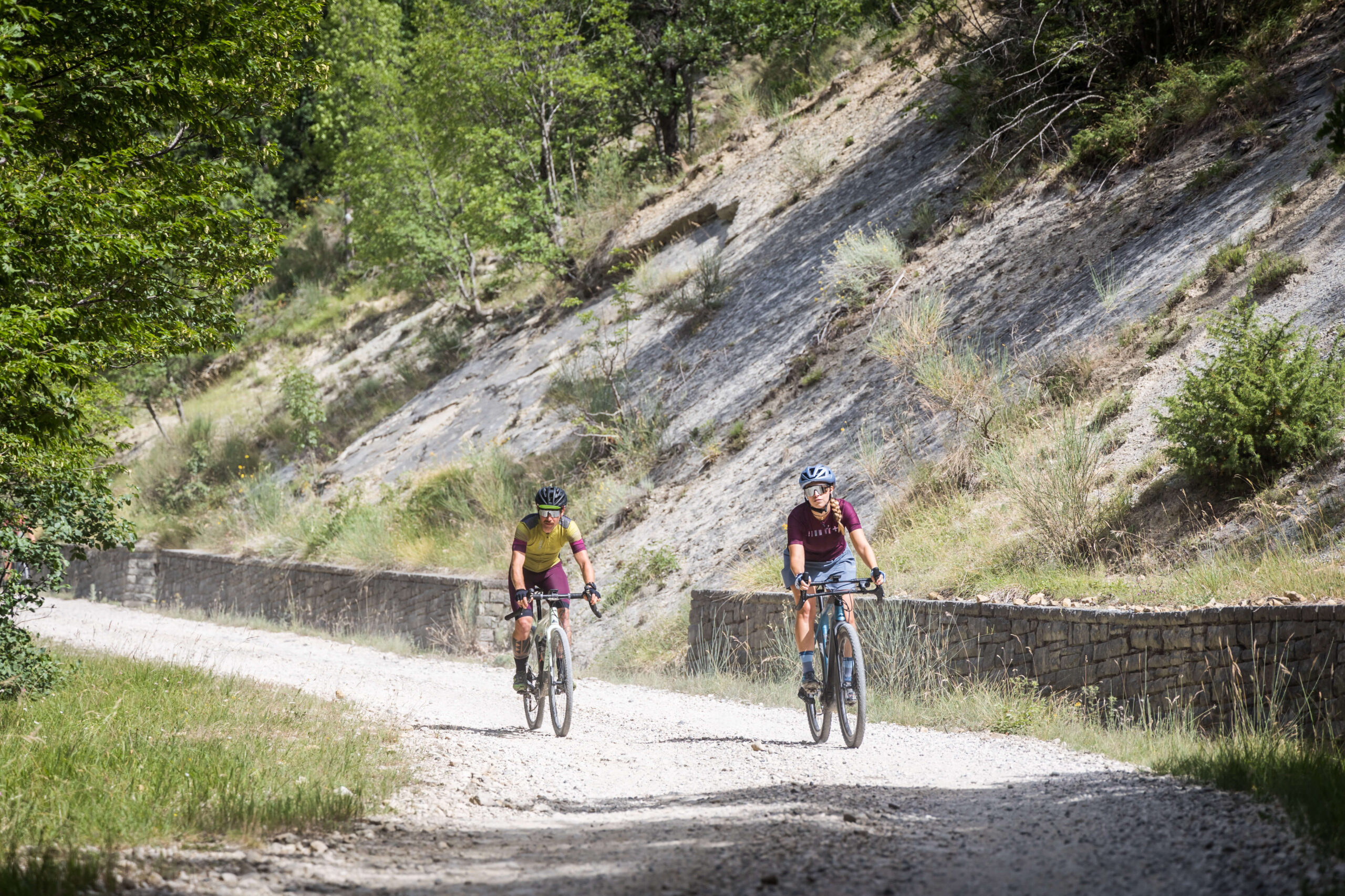 Due ciclisti gravel pedalano su strada bianca tra boschi e pareti rocciose durante Unpaved Roads.